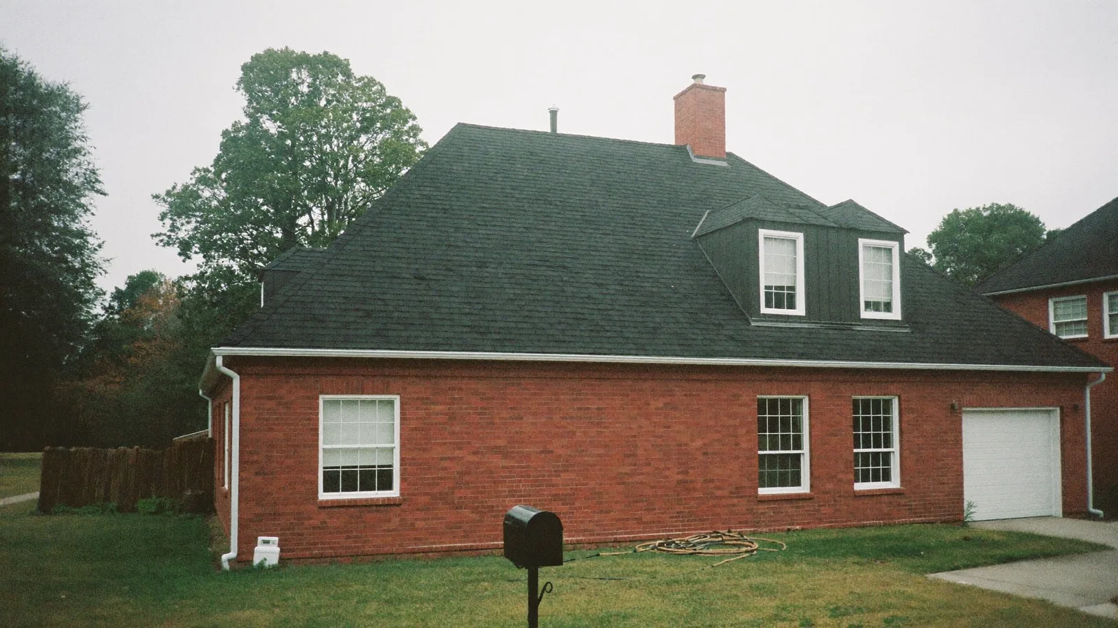 A red brick house with a dark architectural shingle roof, twin dormers and a brick chimney, under flat overcast light