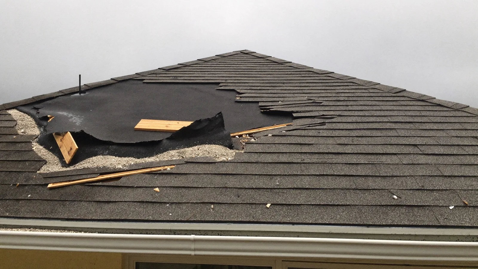 A hip roof with a large section of shingles torn away, exposing black underlayment, bare sheathing and splintered lumber scattered across the slope